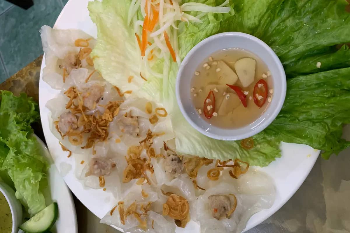 A plate of white rose dumplings topped with fried shallots, with a dipping sauce and fresh greens