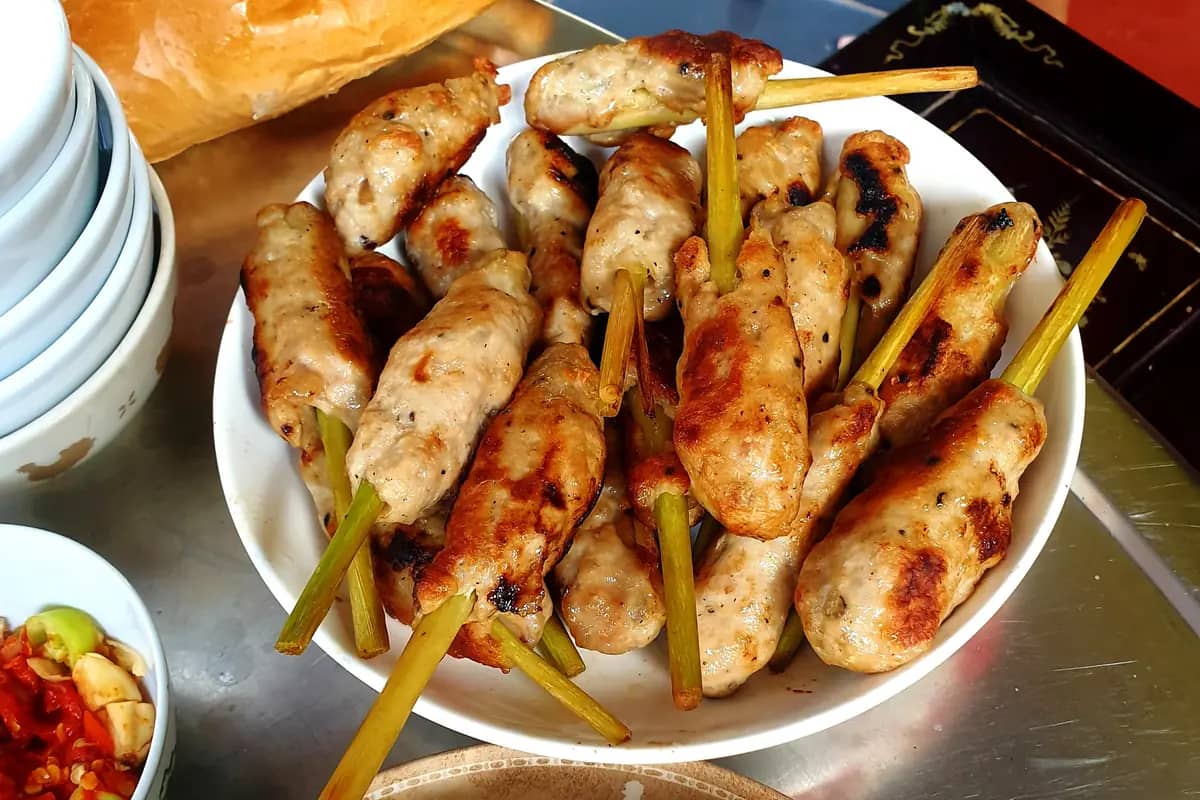 Pork grilled on lemongrass skewers piled in a bowl, a chilli dip and bread beside it