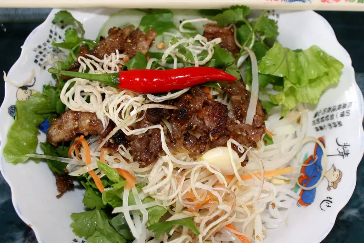 A plate of bun thit nuong: rice noodles under grilled pork, lettuce, herbs and a red chilli
