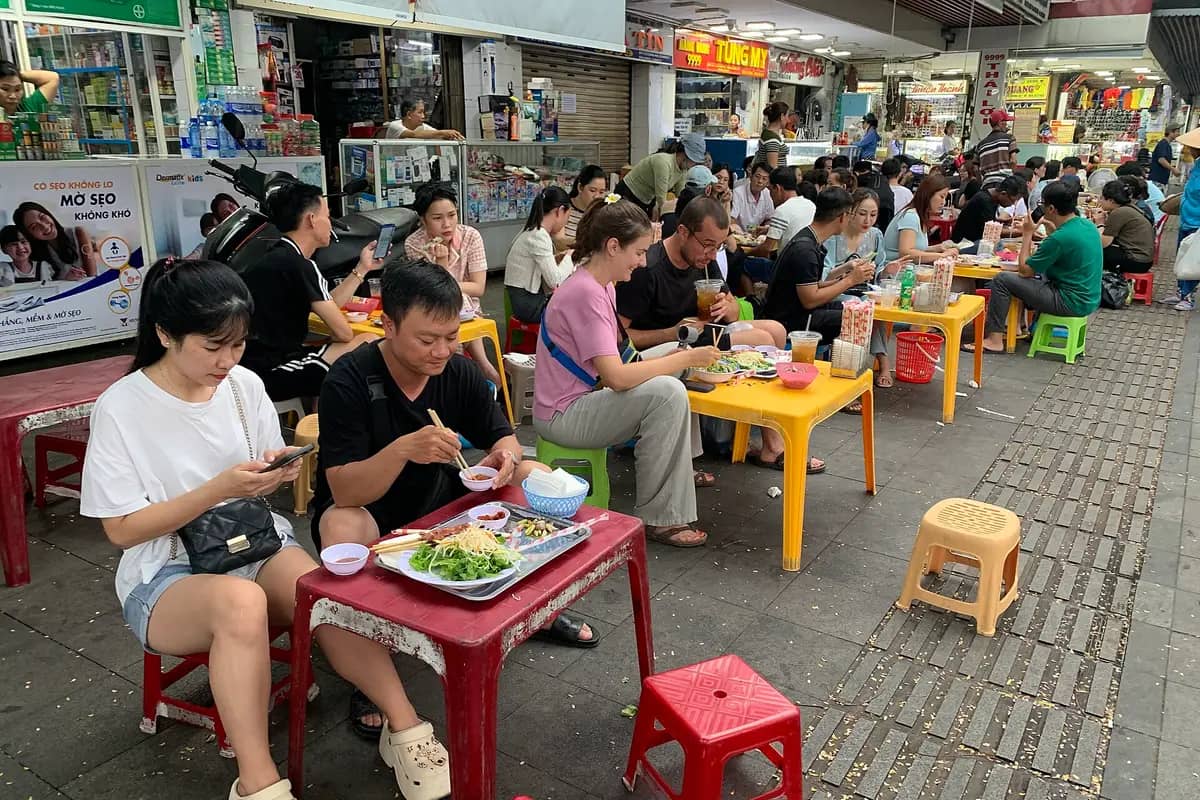People eating at low tables and plastic stools by the food stalls of Dong Ba Market