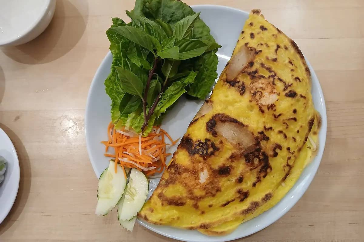 A banh xeo pancake folded on a plate with herbs, lettuce and pickled carrot