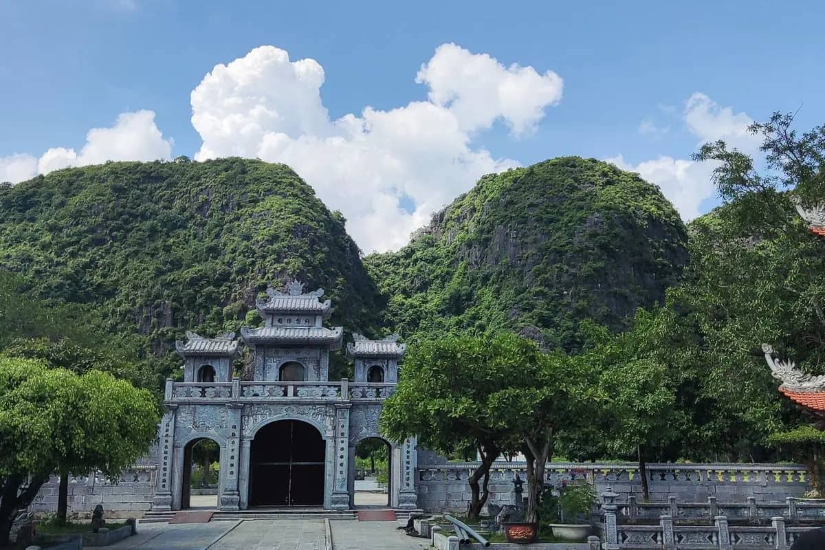The grey stone gate of Thai Vi temple under two rounded karst hills, trees in the courtyard