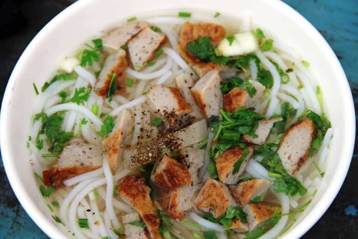 A bowl of bun cha ca: rice noodles with sliced fishcake, herbs and black pepper in a clear broth