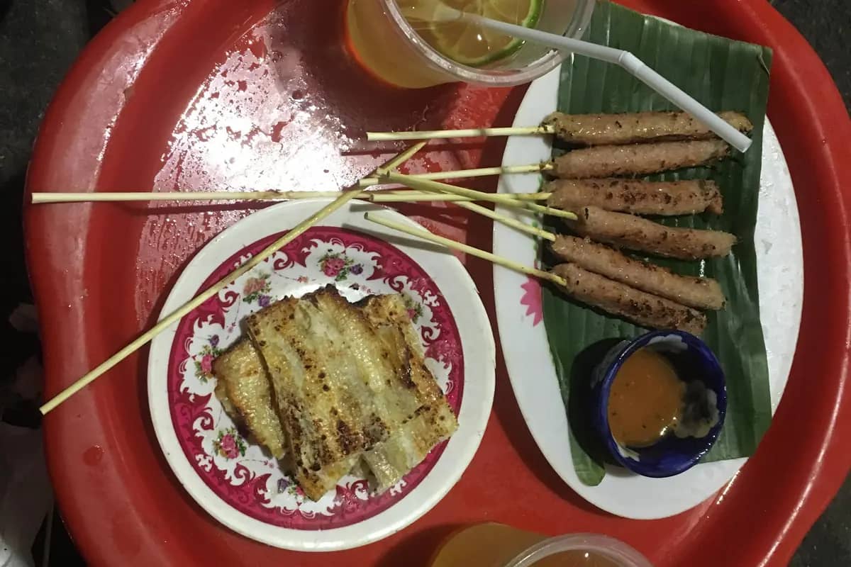 Street food on a red tray: grilled nem nuong pork skewers, a dipping sauce and iced tea