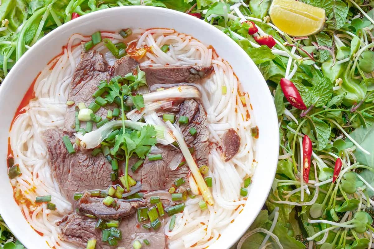 A bowl of beef noodle soup with herbs and chilli, a plate of fresh greens beside it