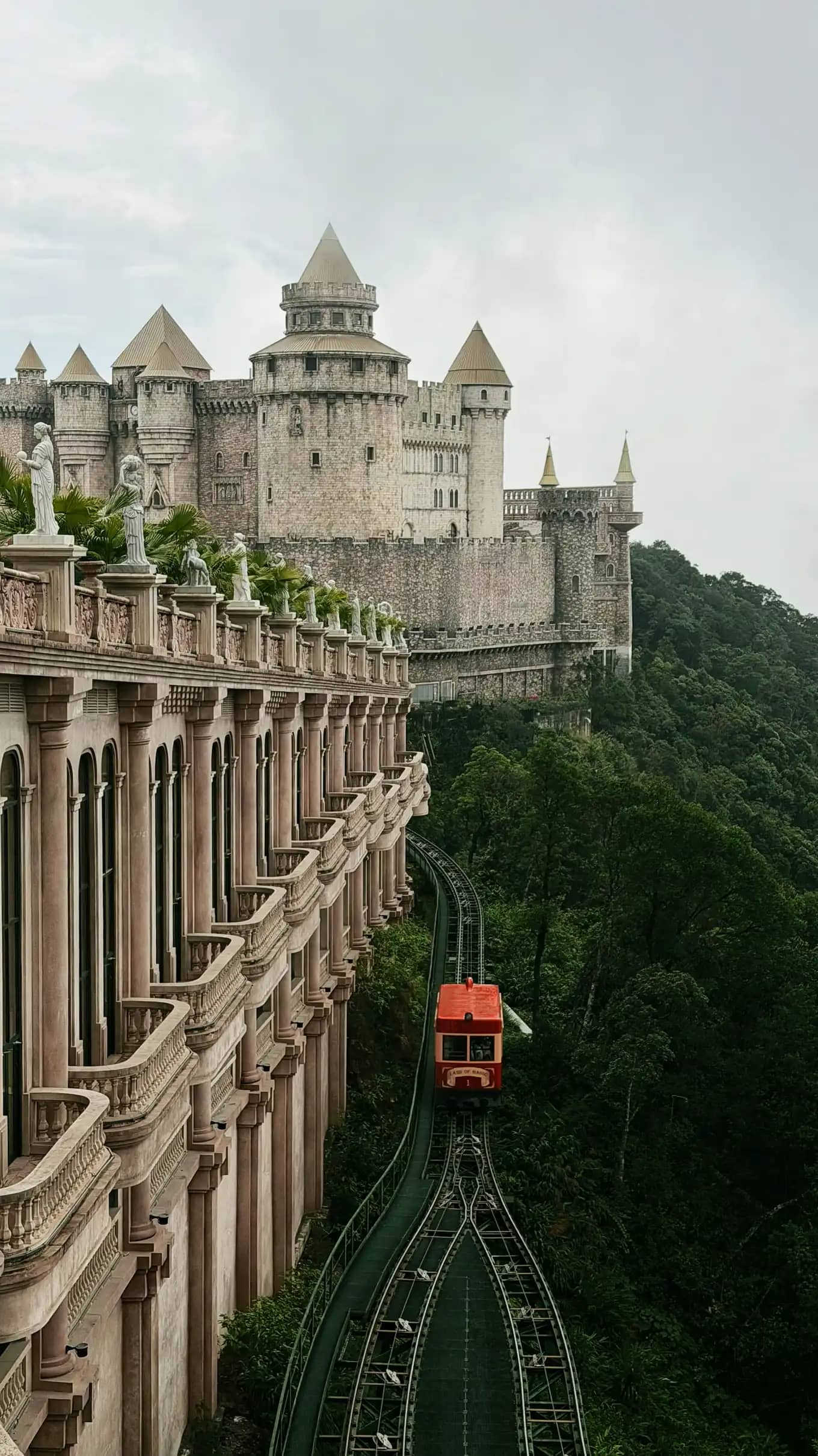 A red funicular train passing a stone castle on the forested slopes of Ba Na Hills