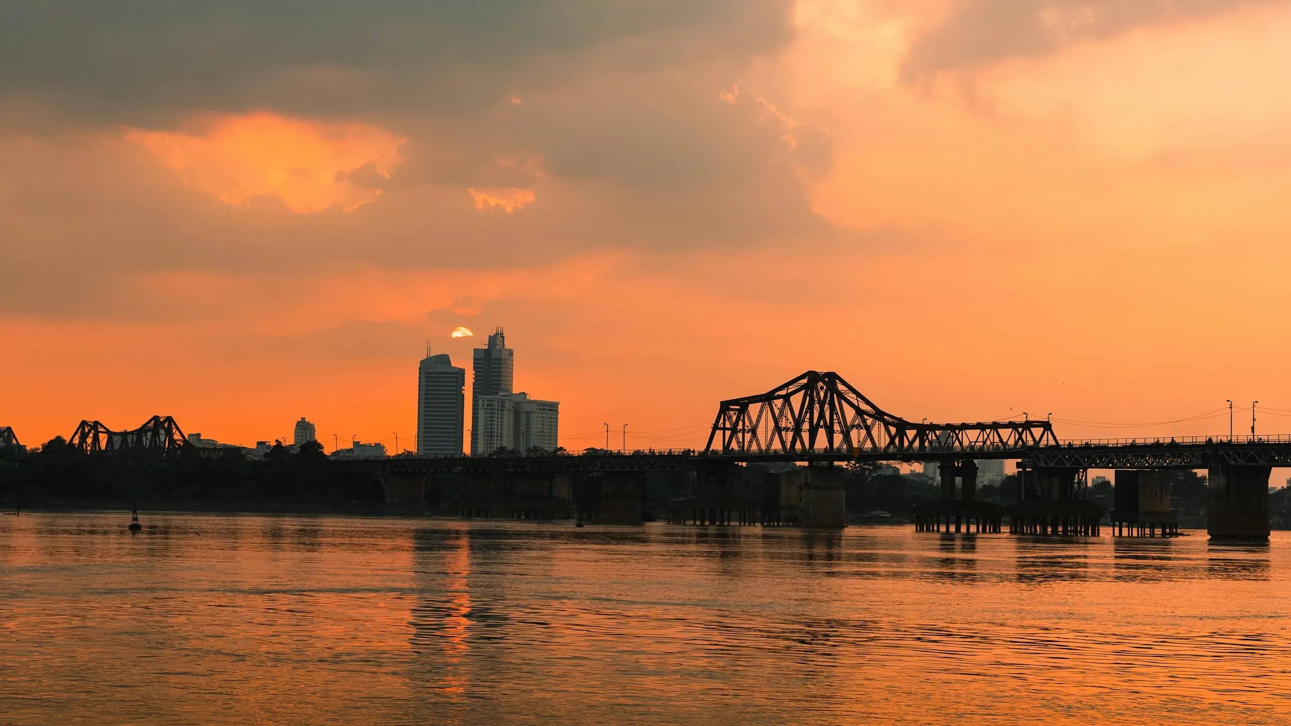Long Bien Bridge and the city's towers in silhouette against an orange sunset sky