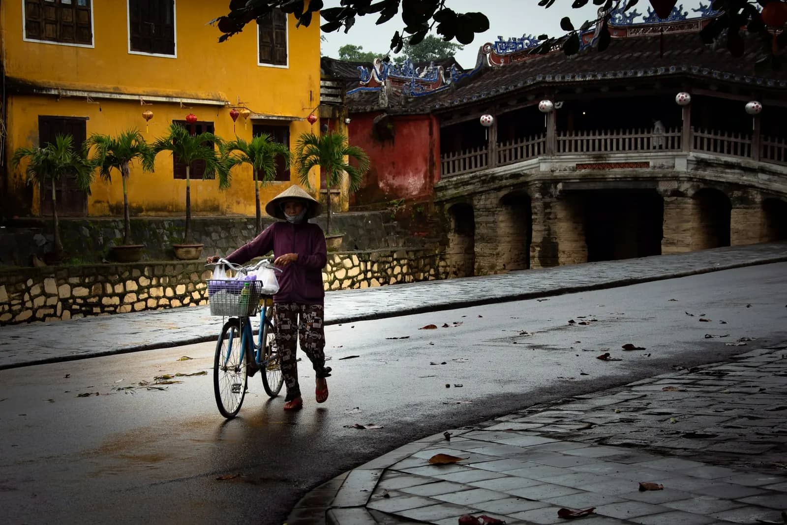 A woman walks her bicycle along a wet street by the Japanese Covered Bridge