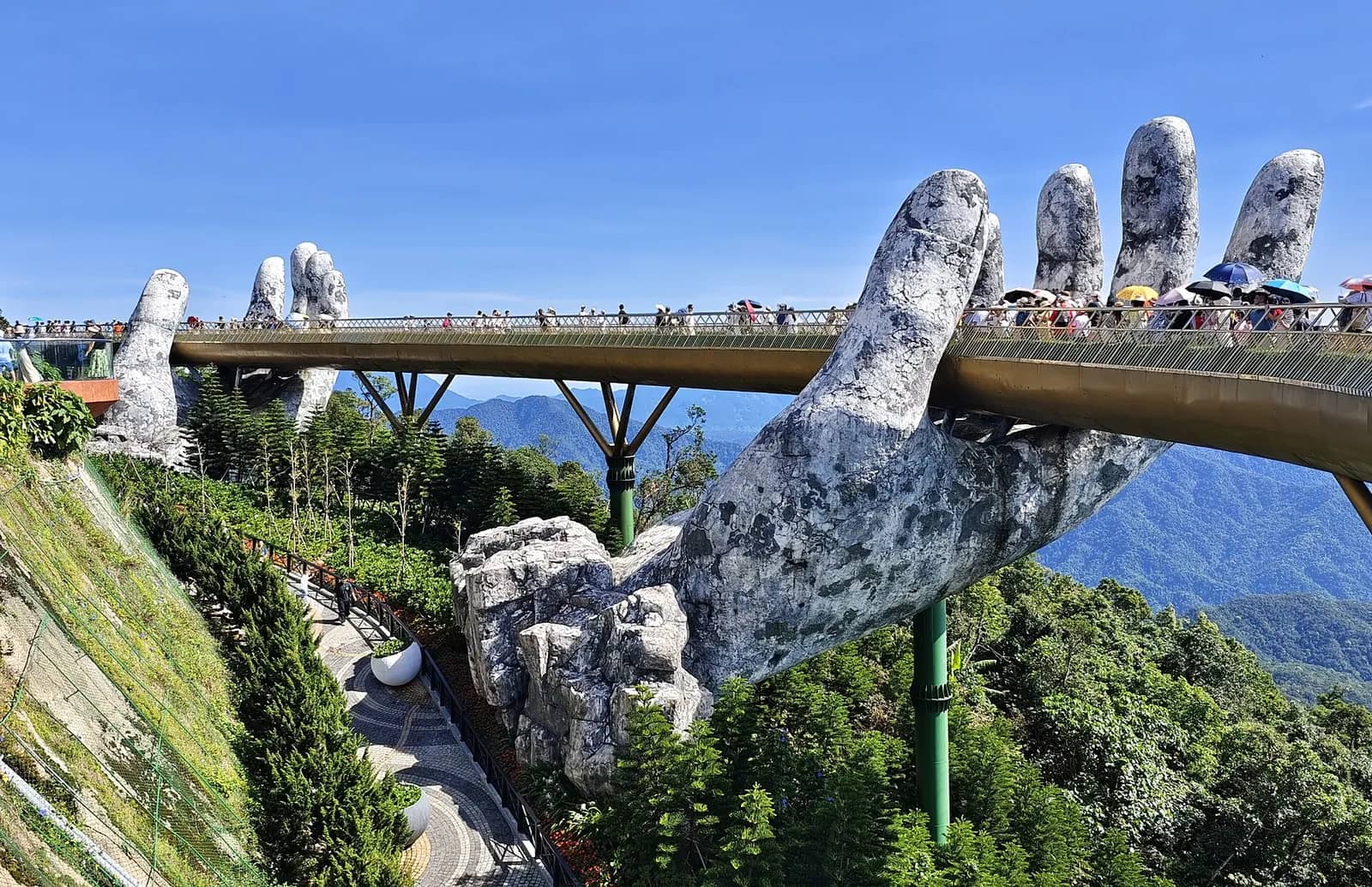 The gold-railed Golden Bridge curving between two giant stone-textured hands above green forest, with mountains behind