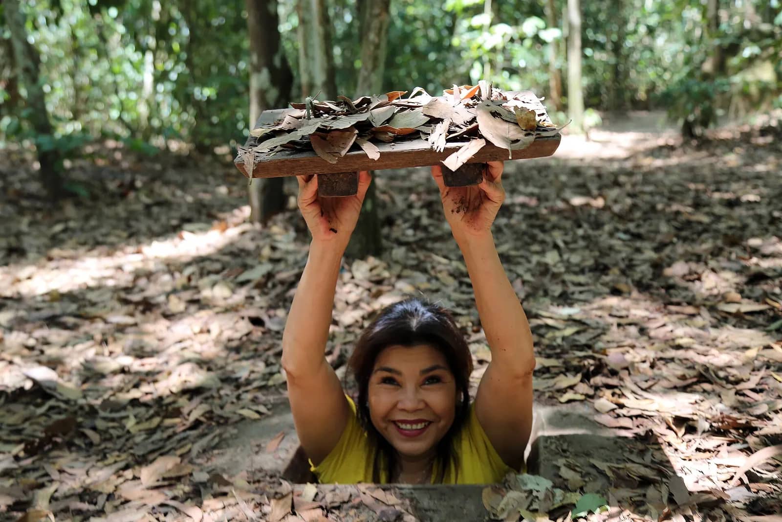 A smiling woman standing in a hidden tunnel hatch, holding its leaf-covered wooden lid above her head in the forest