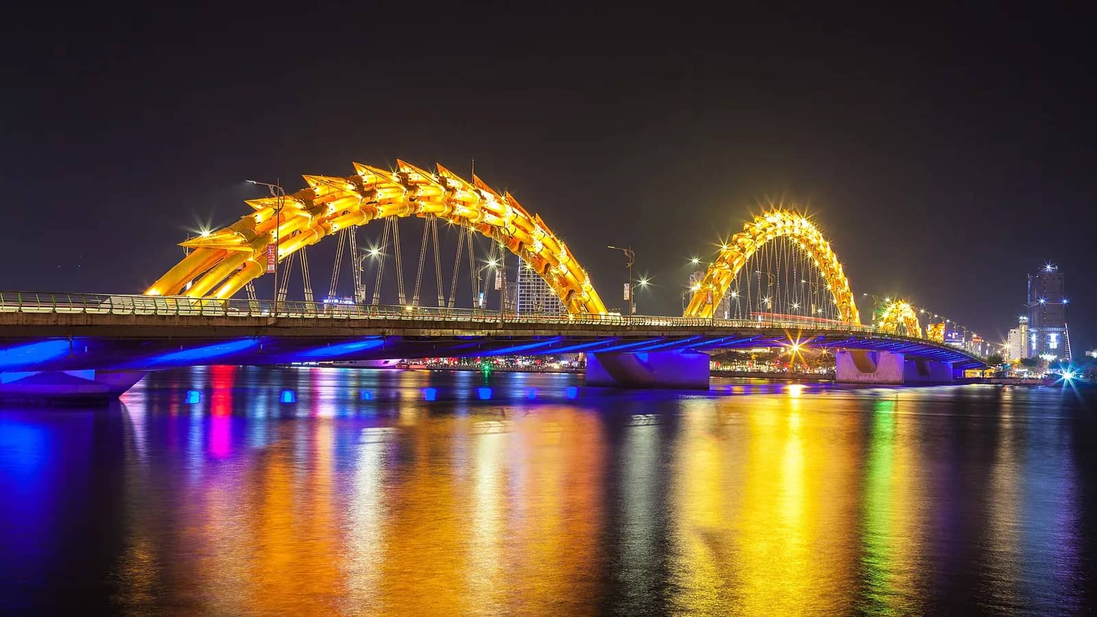 The Dragon Bridge at night, its arched golden dragon body lit up over the Han River with colored reflections in the water