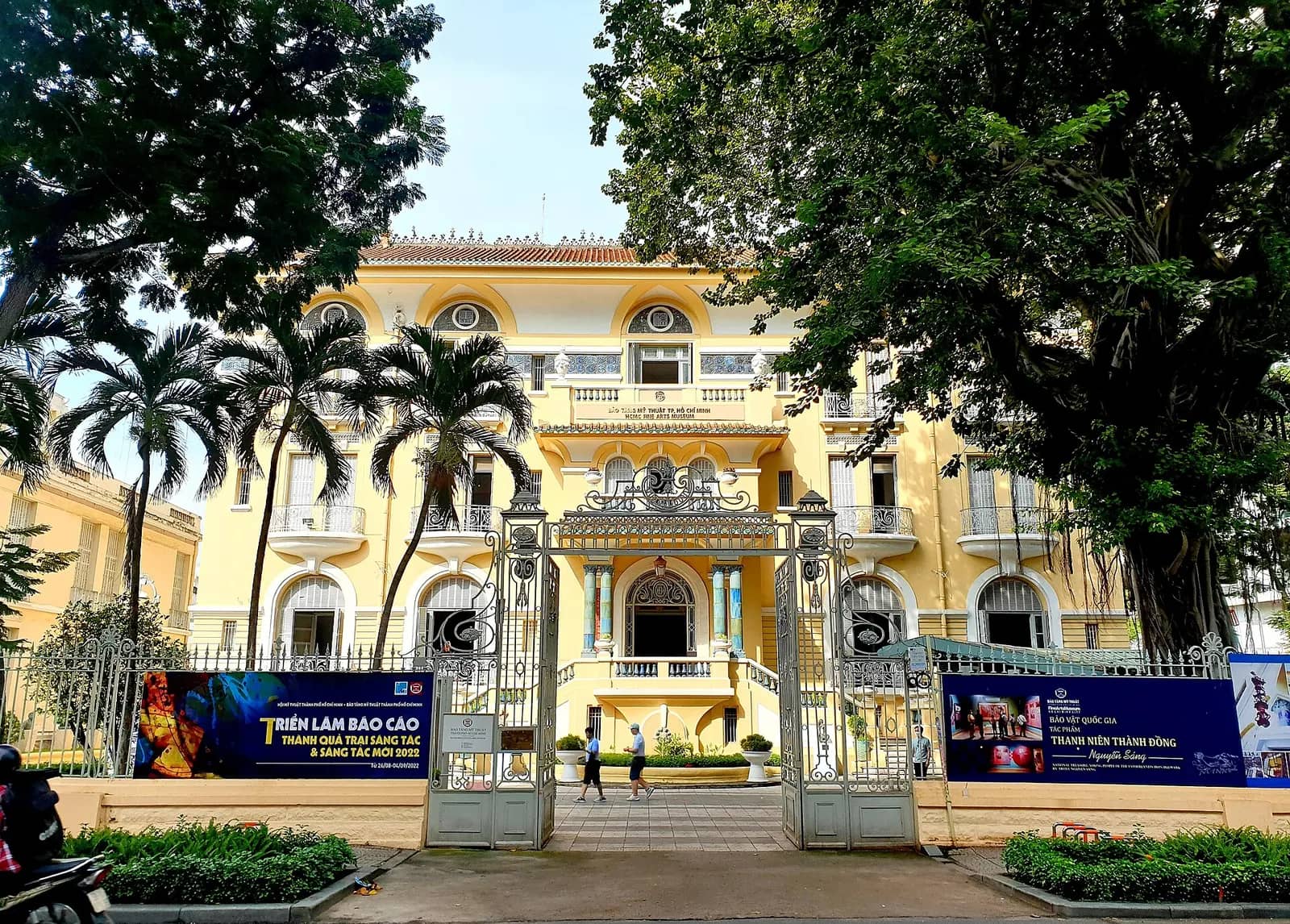 Yellow colonial villa of the Ho Chi Minh City Fine Arts Museum behind an iron gate, with palms and exhibition banners
