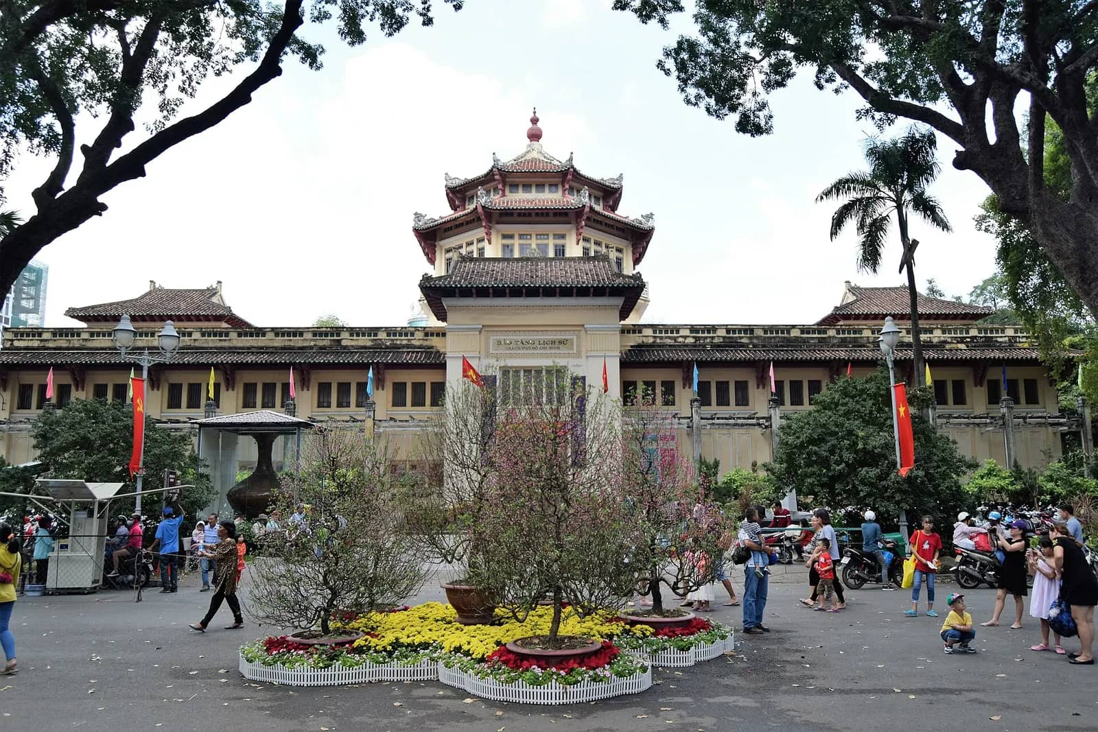 Ho Chi Minh City History Museum, a long yellow building with a tiered pagoda-style tower, flower beds and visitors in front