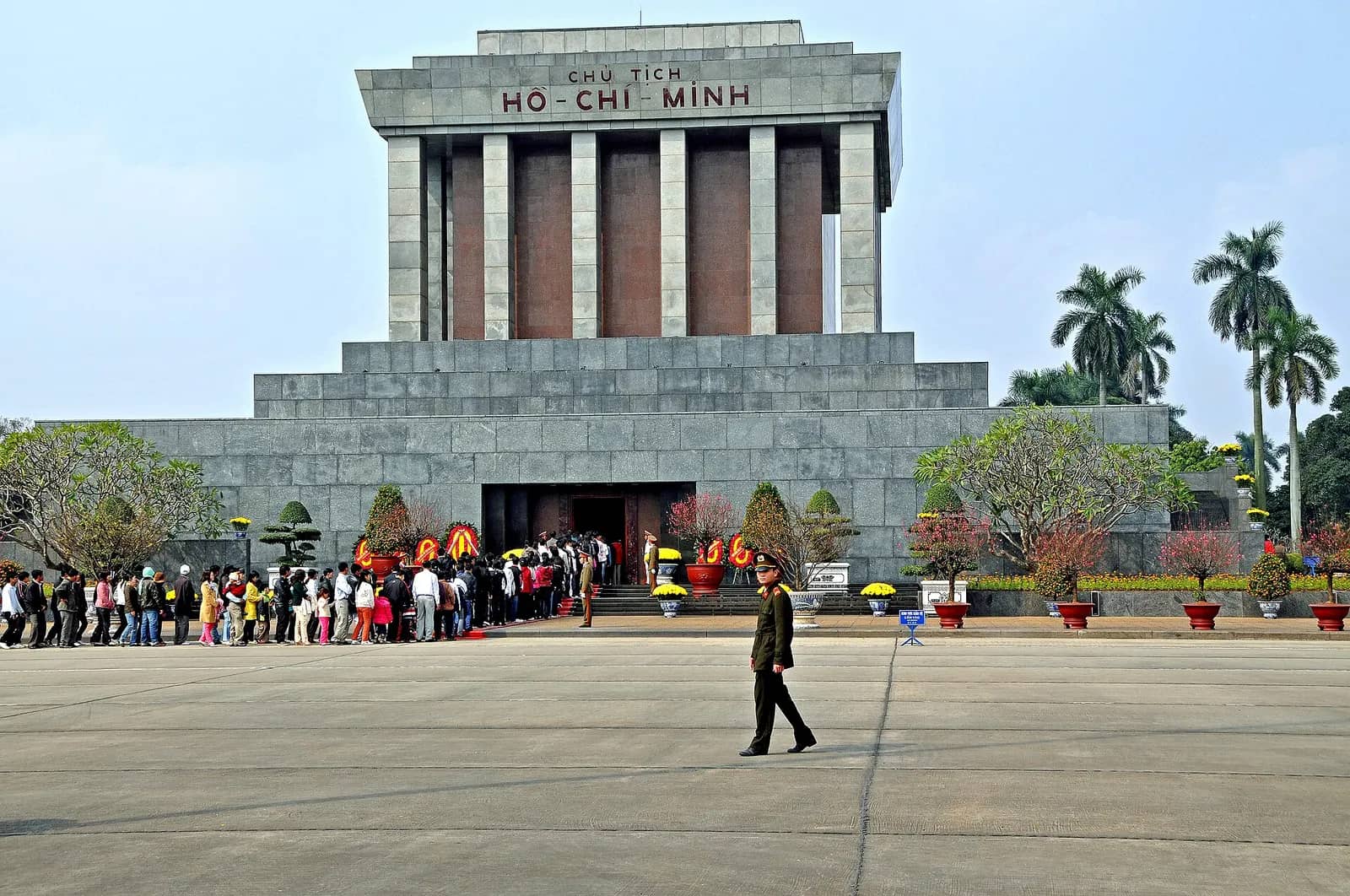 Grey granite Ho Chi Minh Mausoleum with a long line of visitors queuing along its base under a blue sky