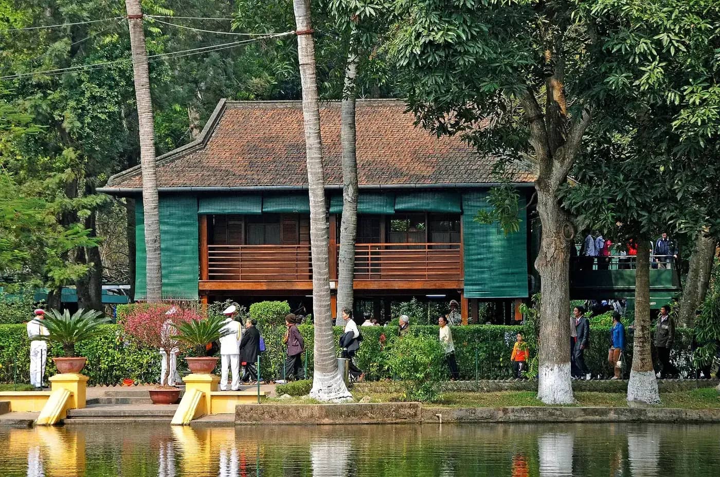 Ho Chi Minh's dark wooden stilt house among tall trees, reflected in a pond, with visitors walking along the bank