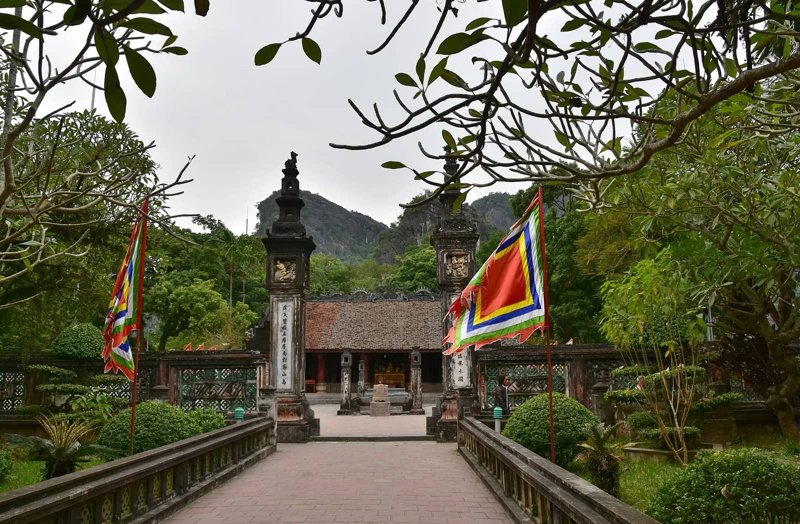 A stone path between flagpoles leading to a pillared temple gate at Hoa Lu, with karst hills behind