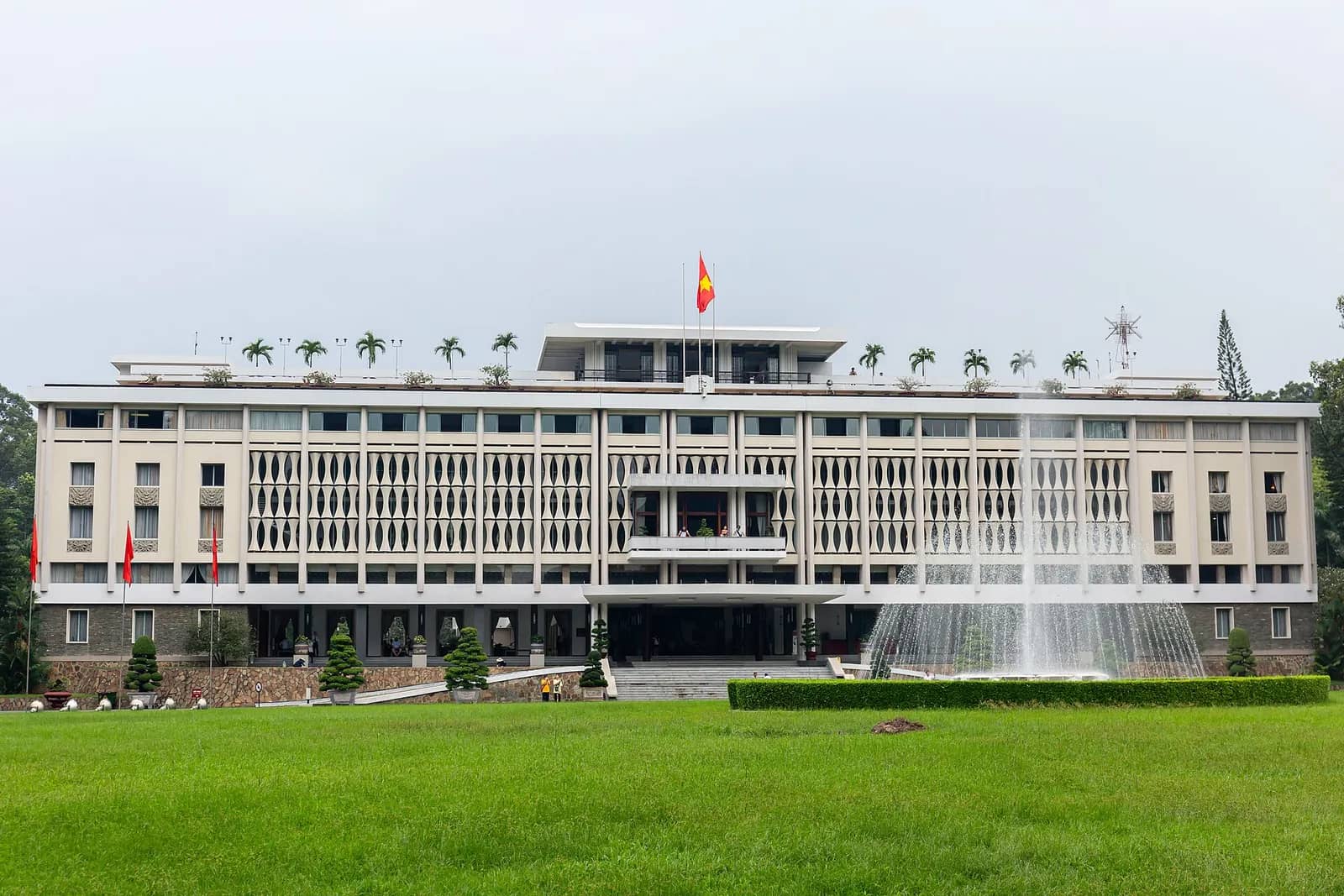 Long white modernist front of Independence Palace behind a wide lawn and fountain, with a red flag on the roof