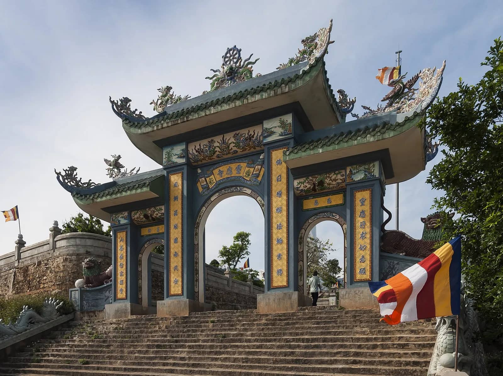 The ornate three-arched gate of Linh Ung Pagoda at the top of a stone stairway, with Buddhist flags and trees on either side