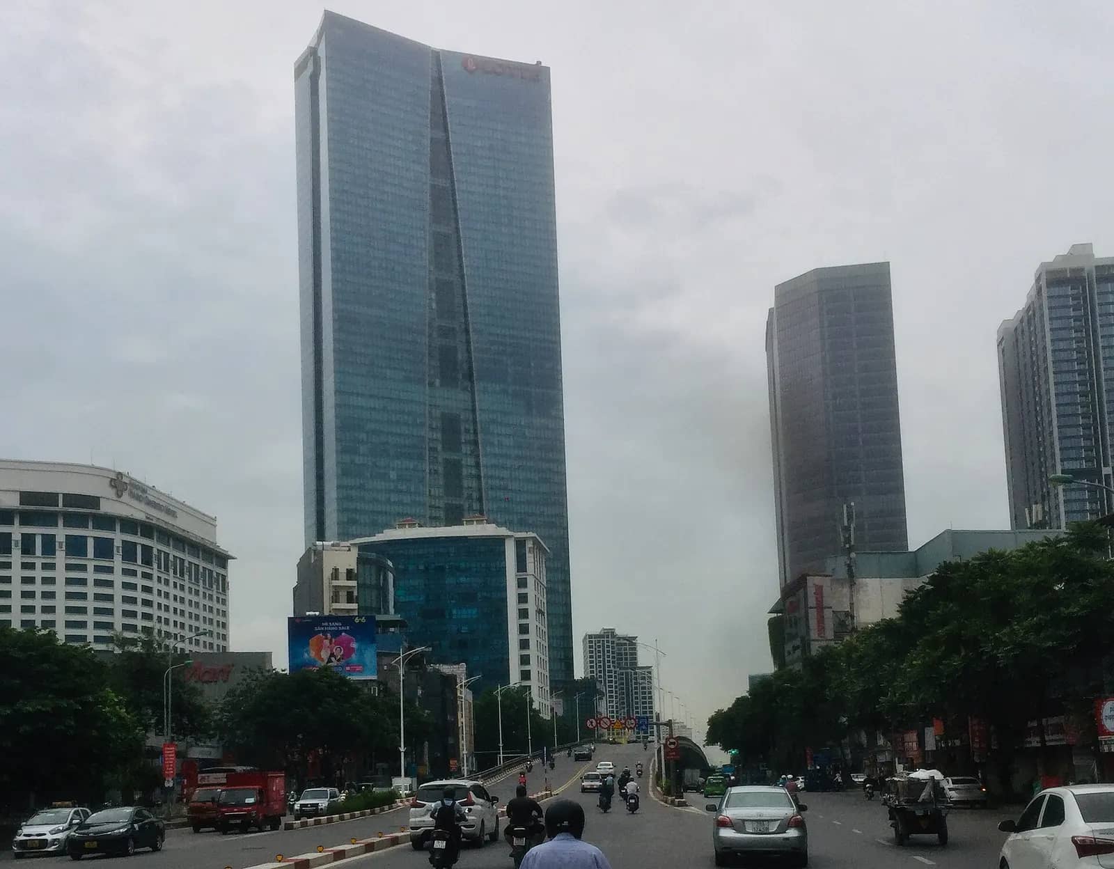 The glass LOTTE Center tower rising above a busy road with motorbikes and lower office blocks under a grey sky