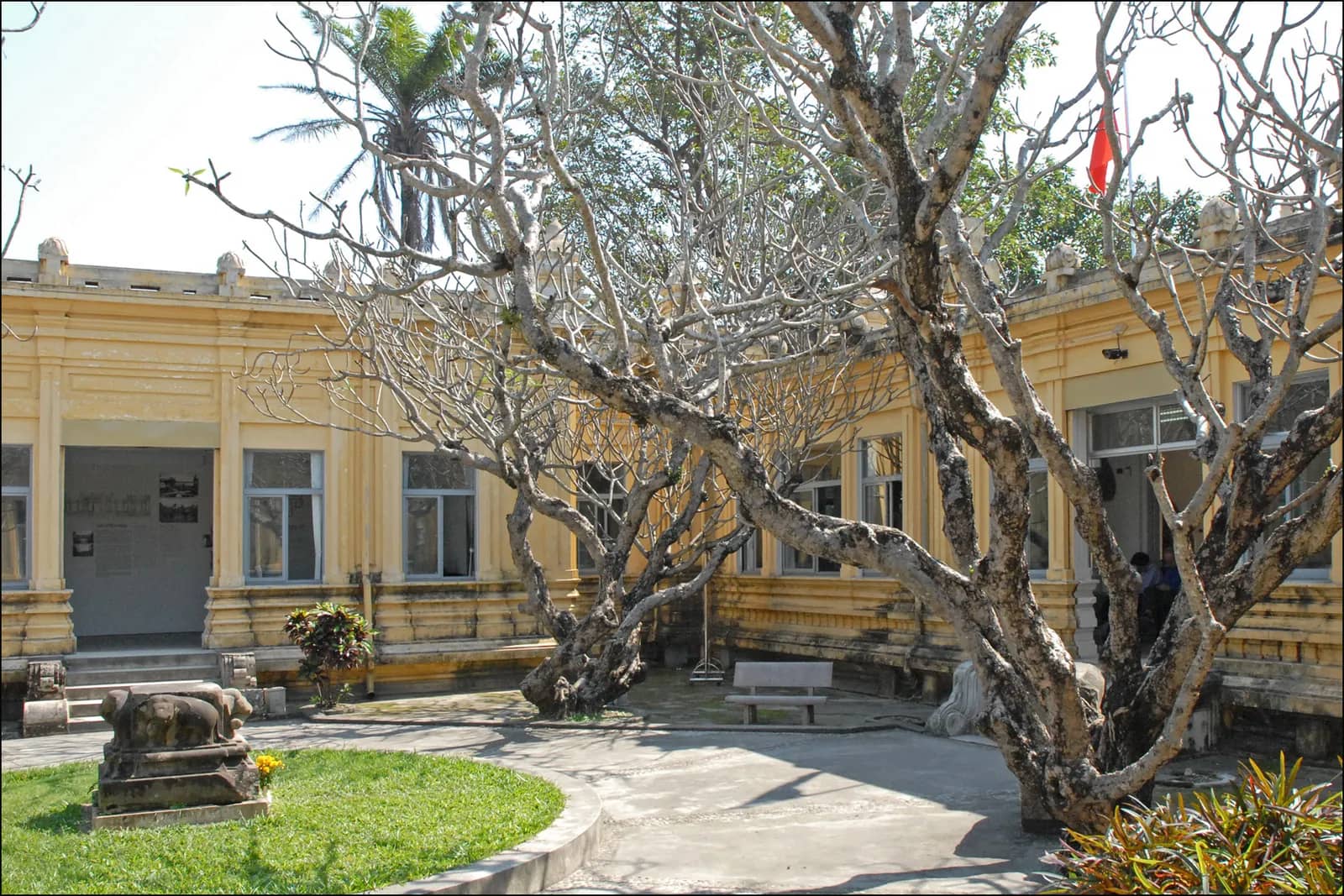 The yellow colonial-era wings of the Museum of Cham Sculpture around a courtyard with frangipani trees and a stone carving on the lawn