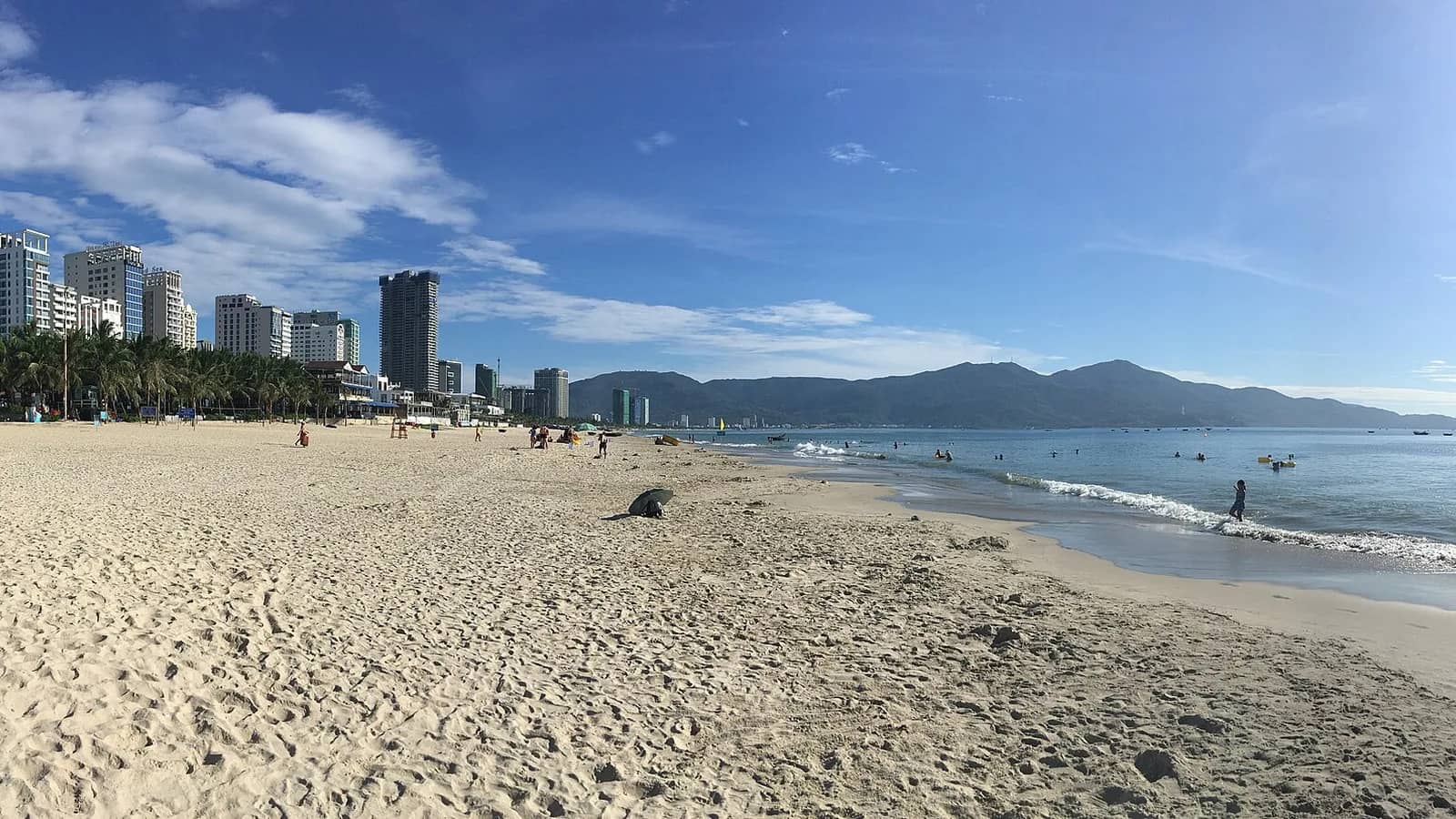 Wide pale sand and gentle surf at My Khe Beach, with hotel towers and palm trees on the left and mountains across the water