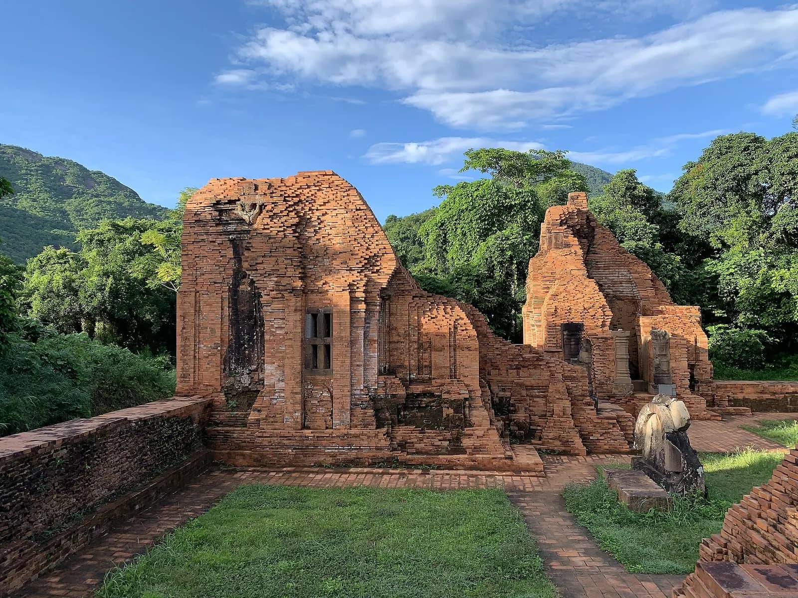 Red brick Cham temple towers of My Son Group A on a lawn, with forested hills behind under a blue sky