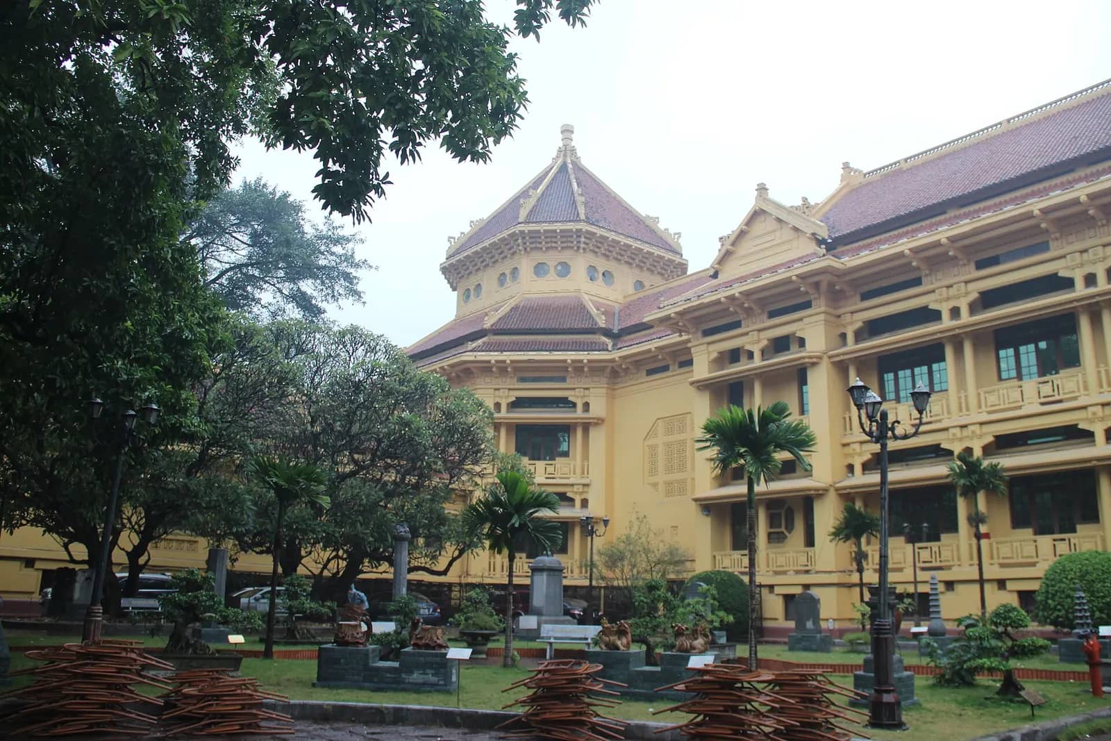 Ochre French colonial museum building with a tiled roof and a domed corner tower, behind palm trees and a garden