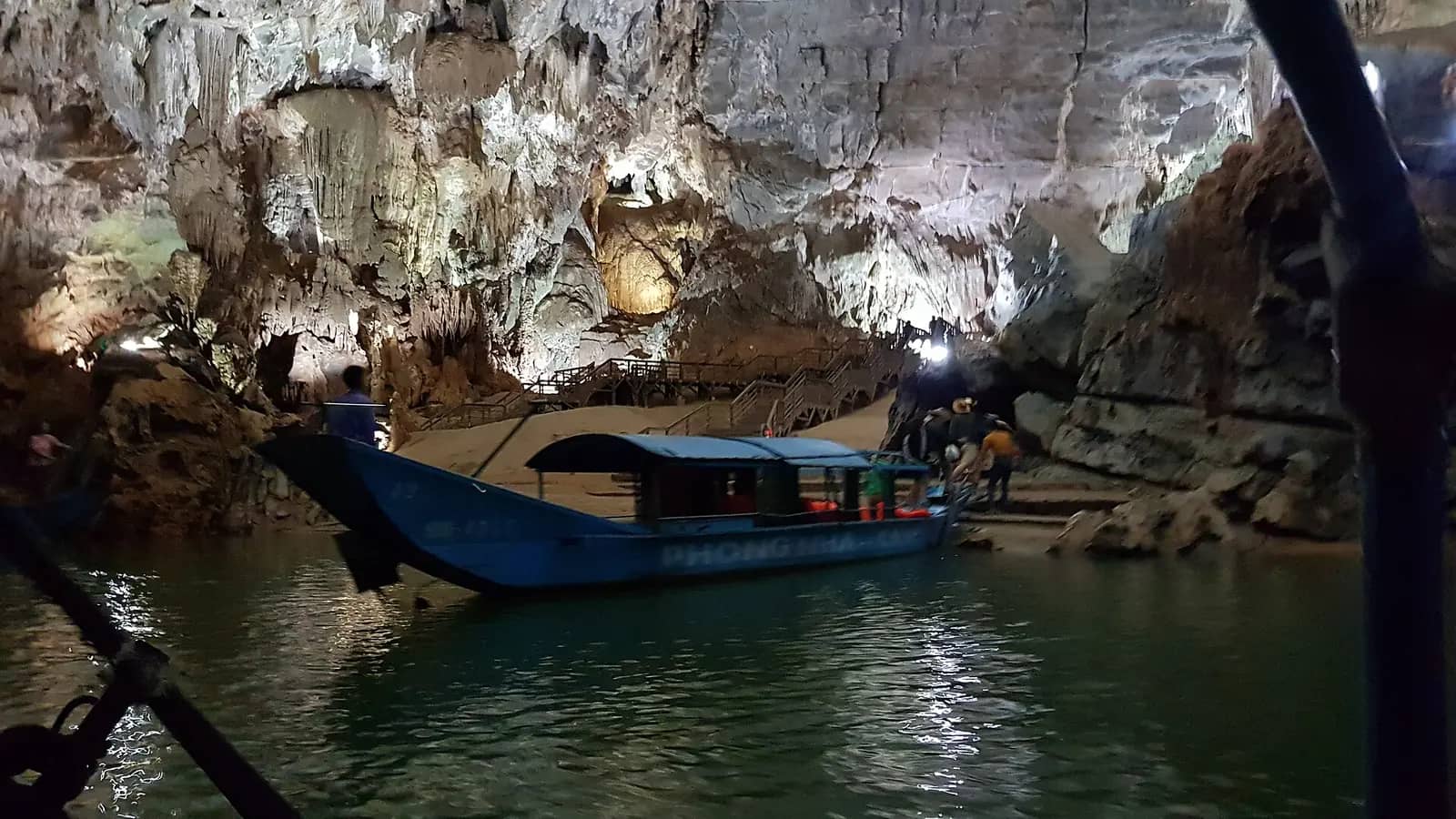 A blue tour boat moored on the green underground river inside Phong Nha Cave, with lit rock walls and a wooden stairway behind