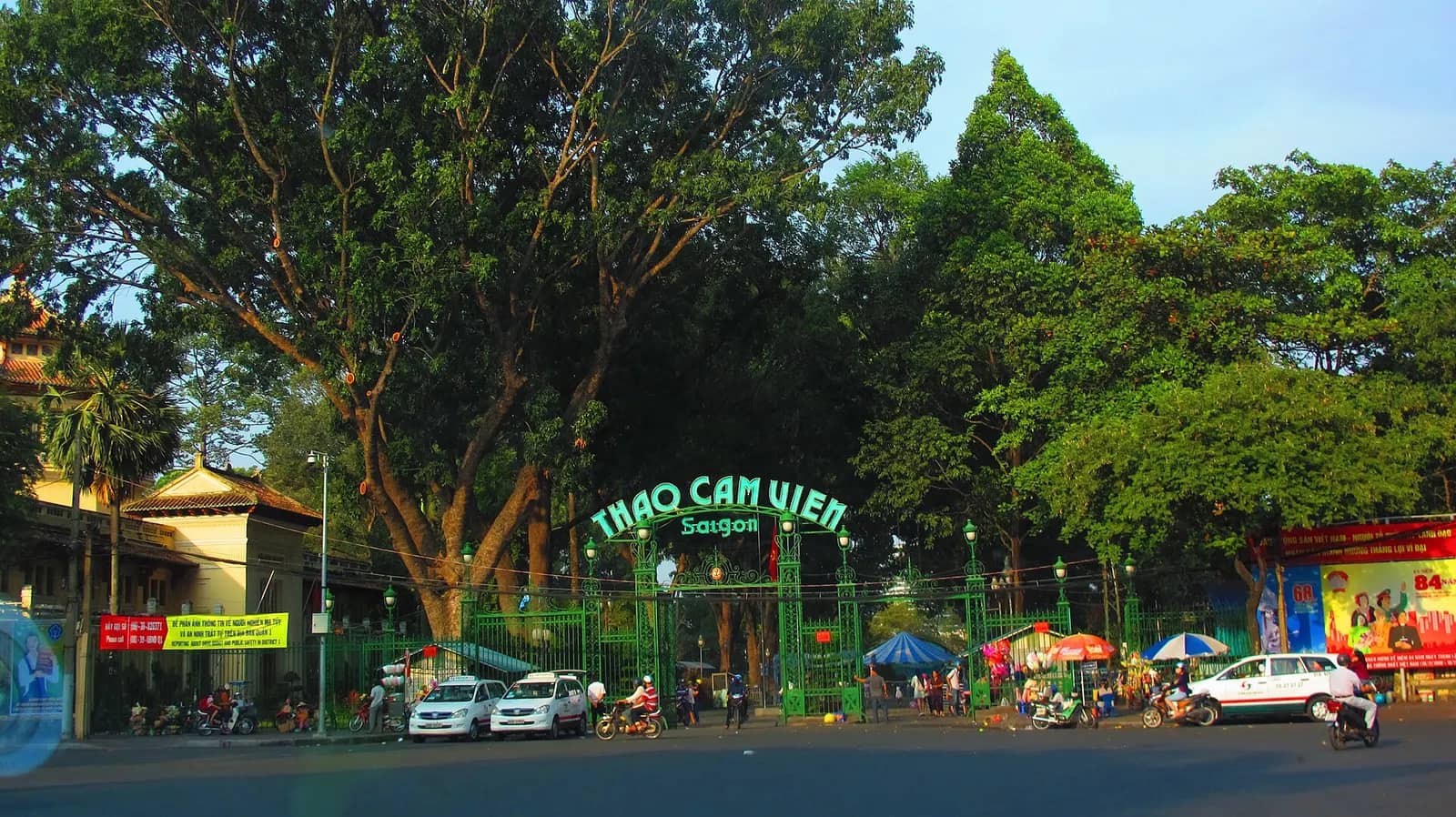 Green arched Thao Cam Vien Saigon gate under tall trees, with cars and motorbikes parked outside
