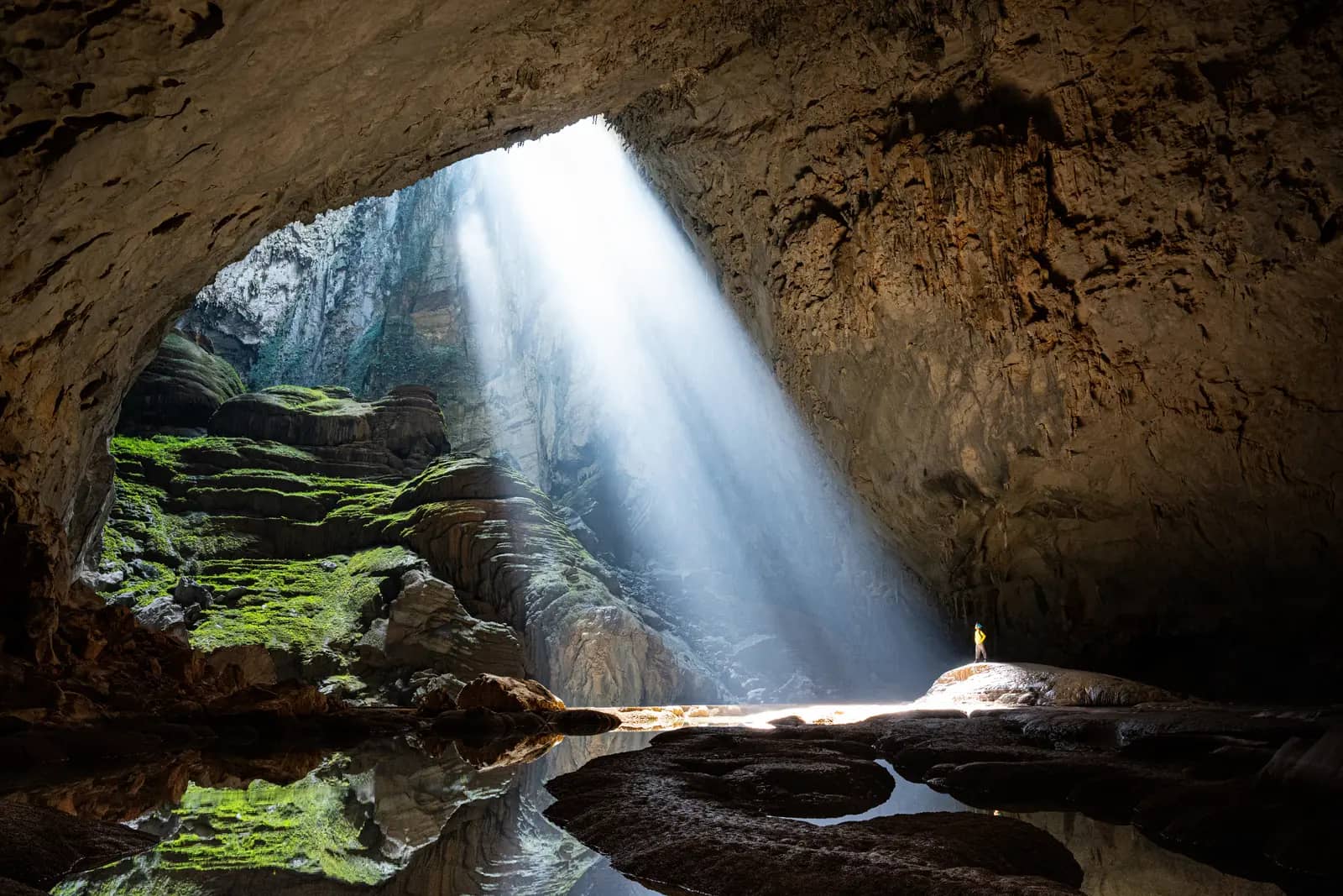 A beam of winter sunlight falling through the first doline of Son Doong onto mossy rocks, with a lone figure standing in the light