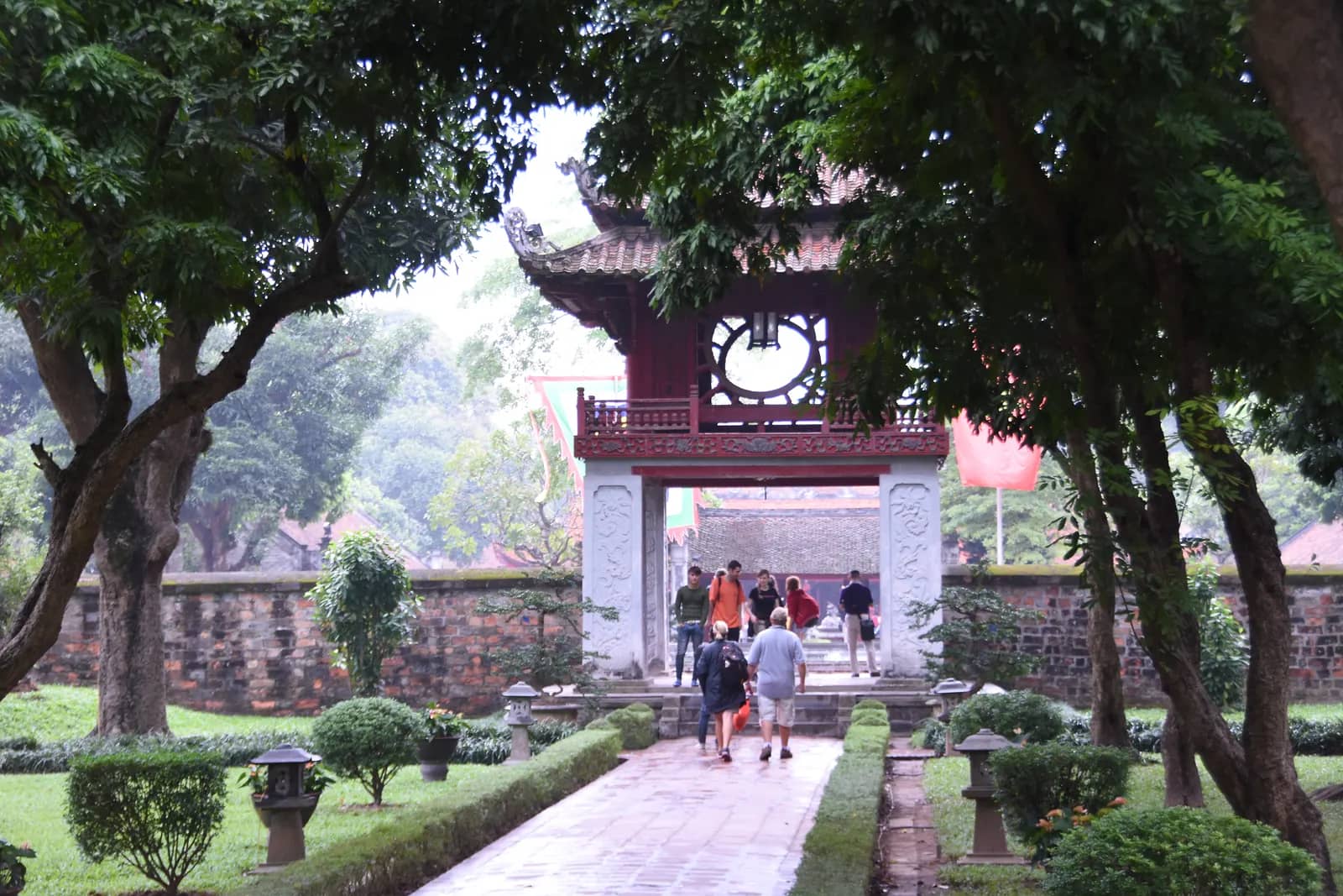 Khue Van Pavilion seen through overhanging trees at the end of a paved path, with visitors walking toward it