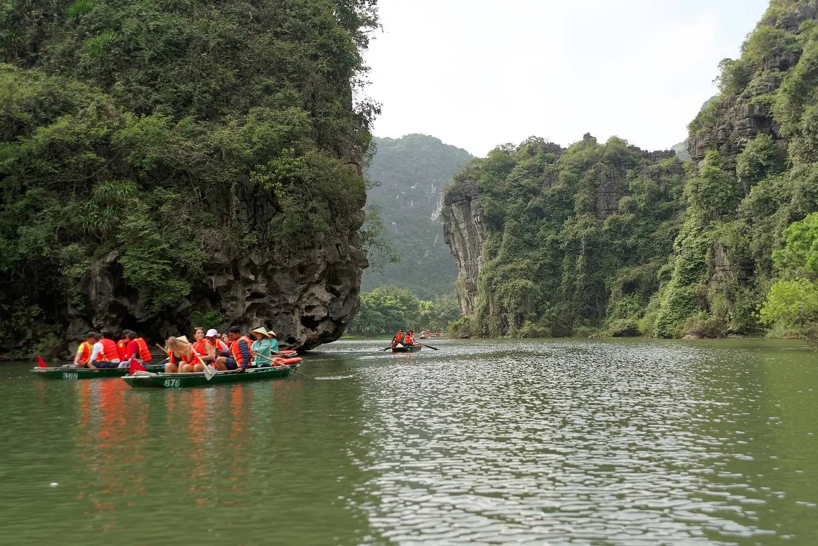 Rowing boats with visitors in orange life jackets crossing open water between forested limestone cliffs at Trang An