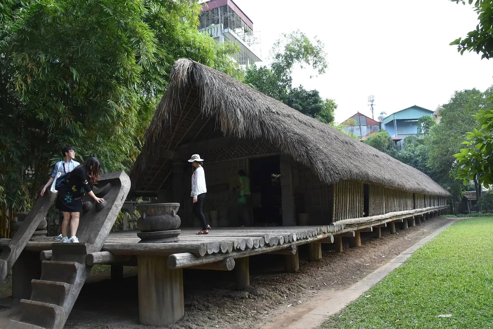 A long Ede stilt house with a thatched roof and a carved log staircase, set among trees in the museum garden