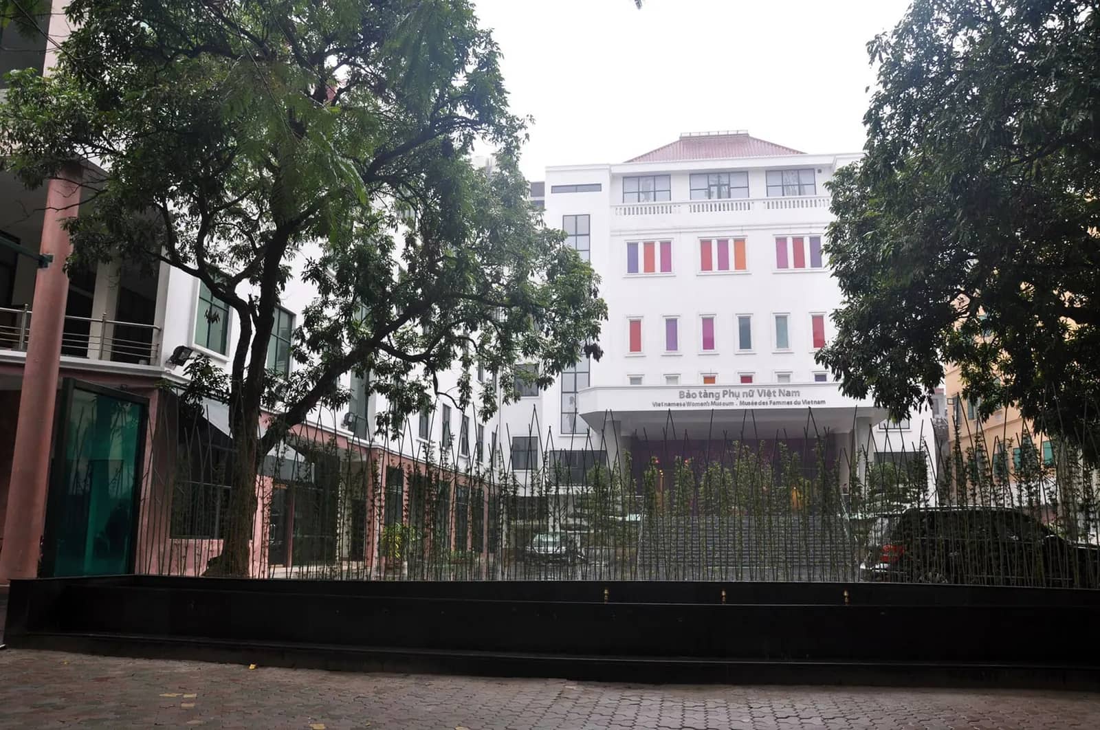 White multi-storey Vietnamese Women's Museum with coloured window panels, behind a fence of thin metal rods and two large trees