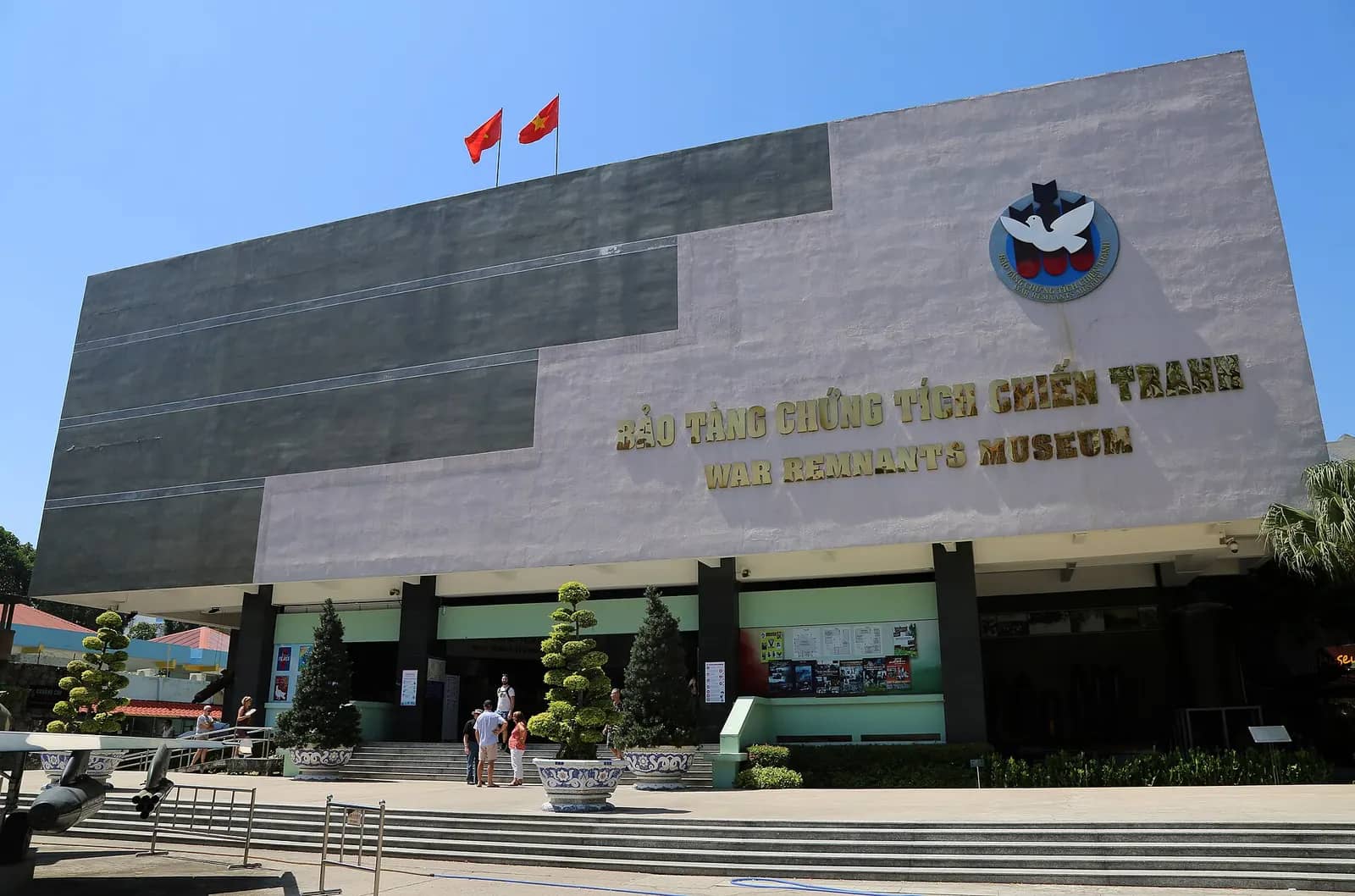 Grey and white front of the War Remnants Museum with its name in Vietnamese and English, a dove logo and two red flags