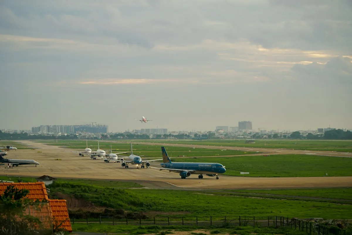 A Vietnam Airlines jet and a line of planes waiting on a city airport taxiway in Vietnam while another jet climbs away under evening cloud