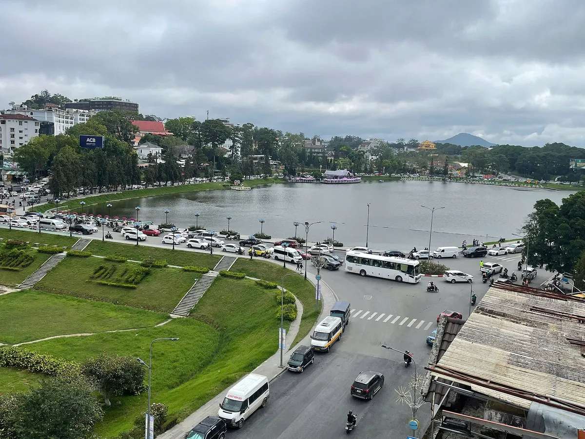 Xuan Huong Lake seen from above in central Da Lat, with a lakeside park, parked cars and buses under grey cloud