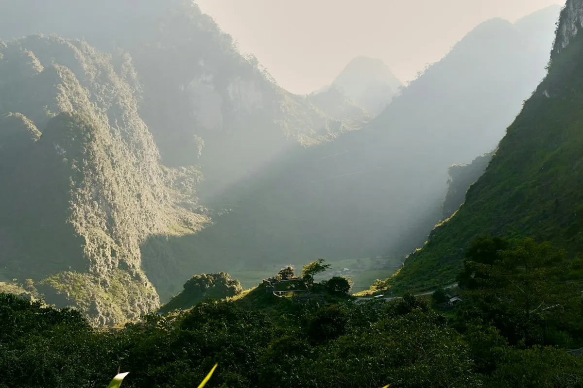 Hazy sunlight over steep karst cliffs and a forested valley toward Duong Thuong village on the Ha Giang Loop