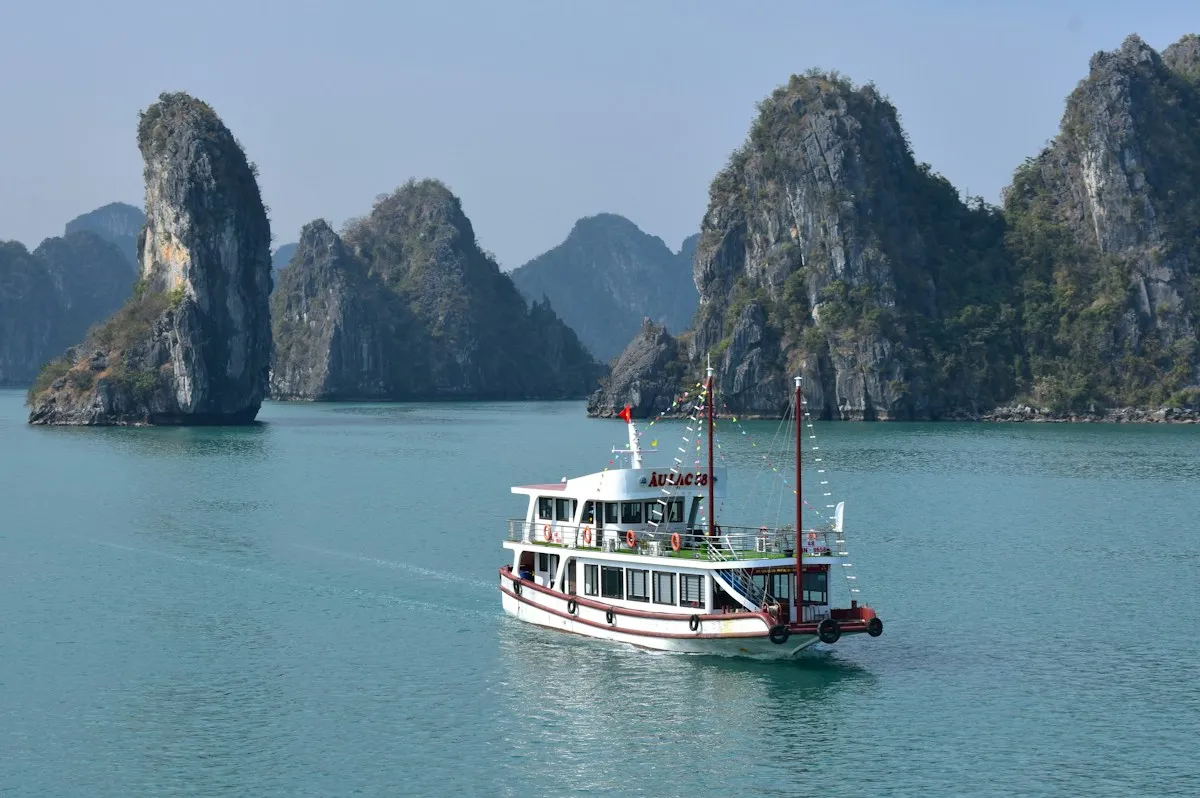 A white sightseeing boat with red flags sailing through calm water past jagged karst peaks in Ha Long Bay