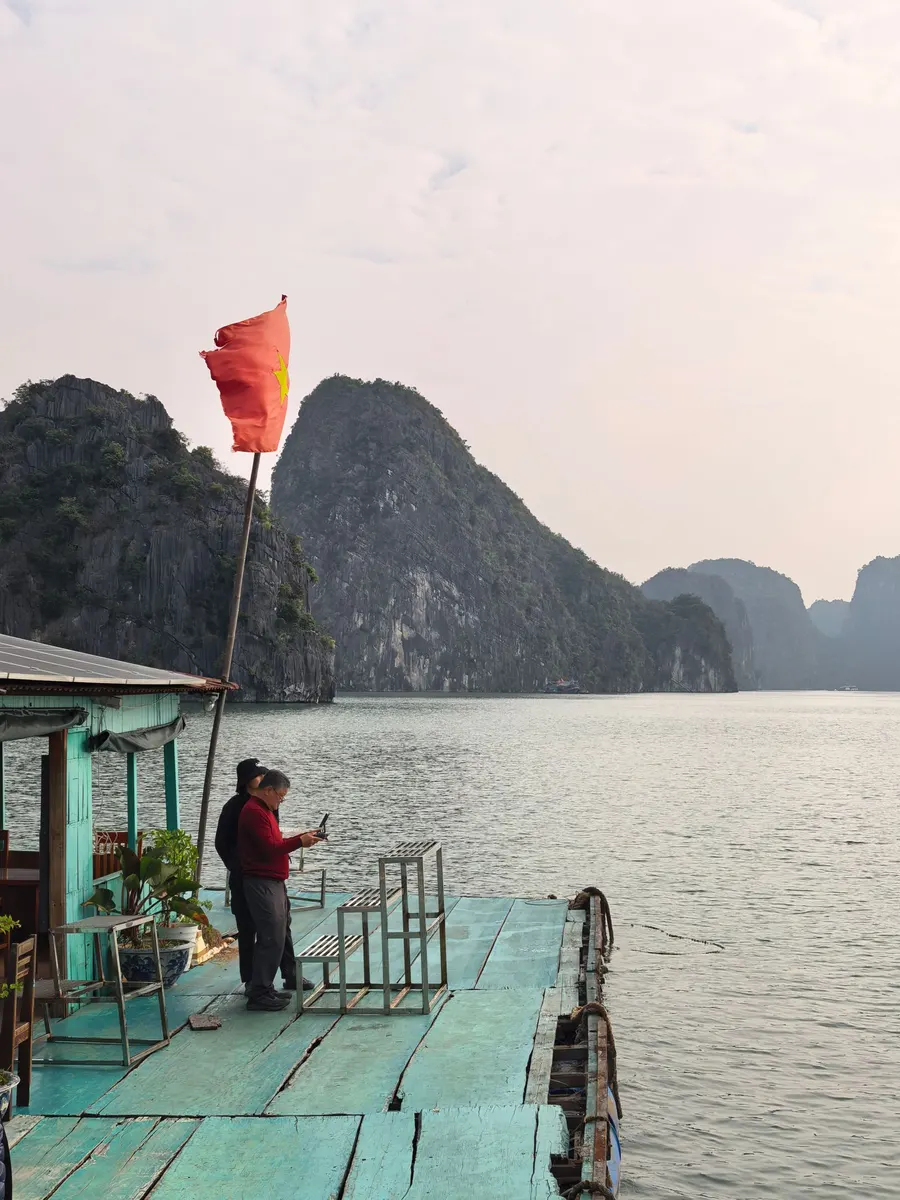 Two people stand on a floating wooden platform under a Vietnamese flag, with limestone islands across the water in Ha Long Bay