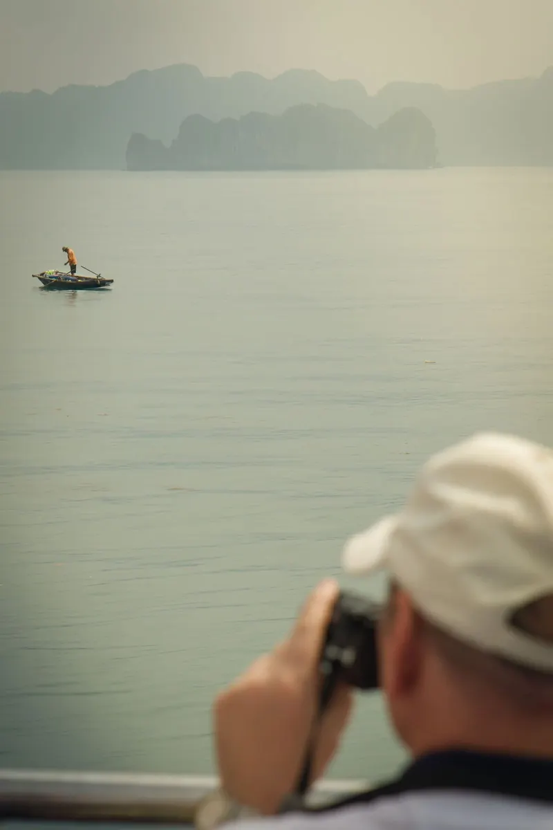 A passenger in a white cap photographs a lone fisherman in a small boat on hazy grey water, karst islands behind
