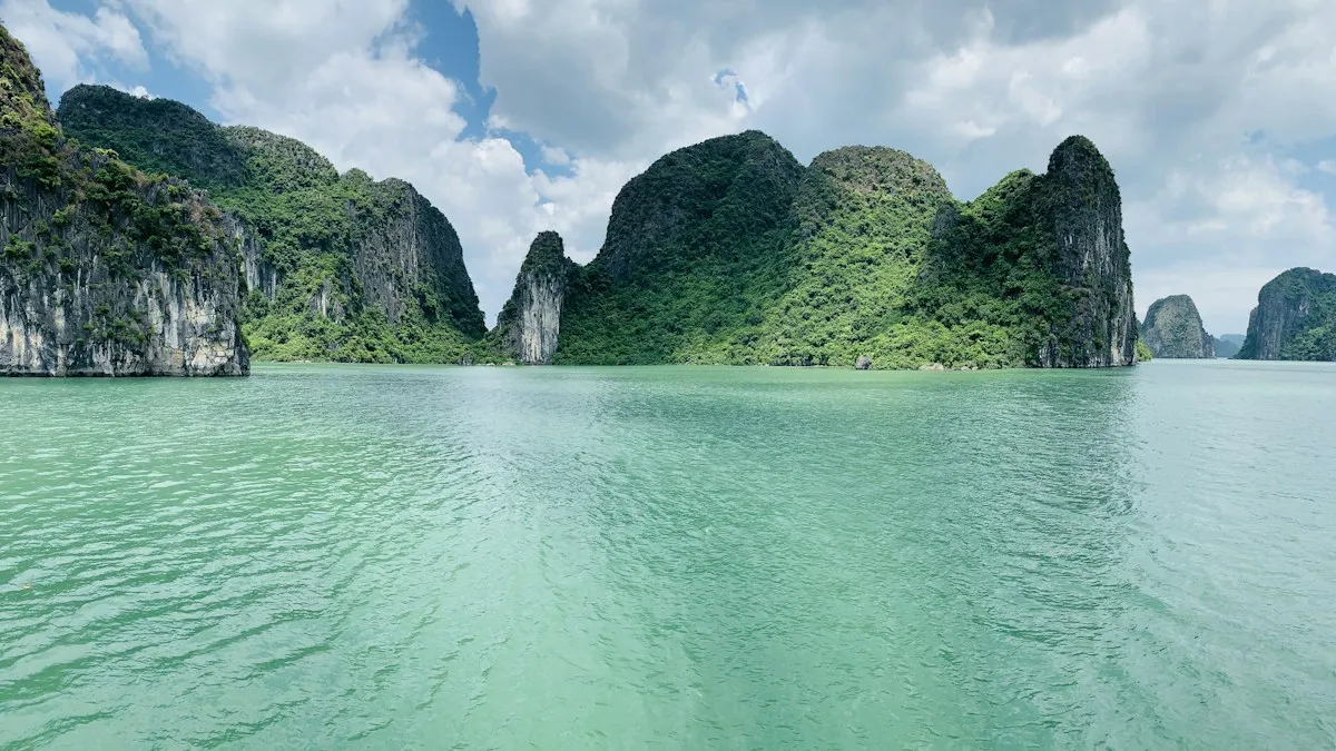 Green karst hills rising from calm jade water in Ha Long Bay under a partly cloudy sky