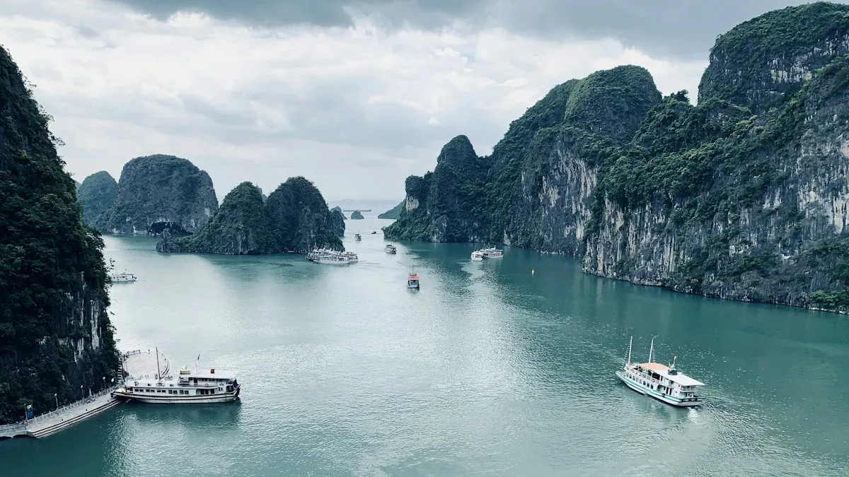 Steep karst cliffs above pale water in Ha Long Bay under a cloudy sky, with two boats near the shore