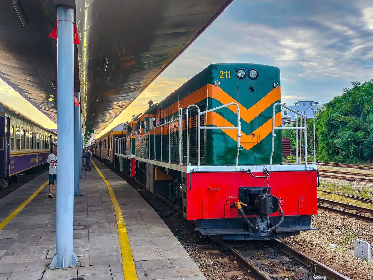 A green and red diesel locomotive at the head of a passenger train beside a covered platform at Hue station