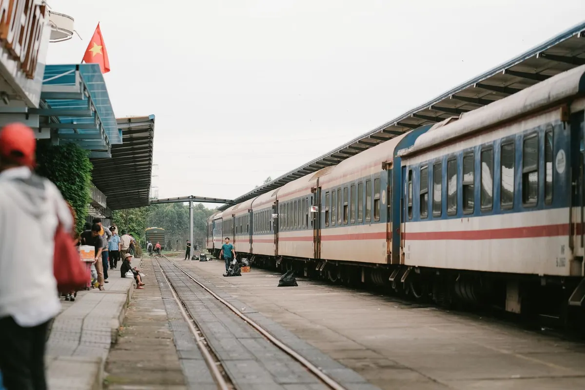 Passengers with bags walking along a station platform beside a long blue and white Vietnam Railways passenger train