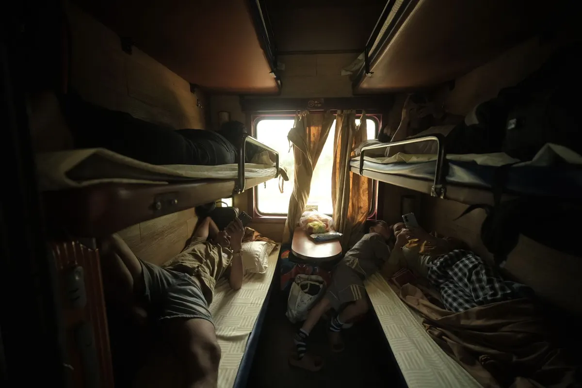 Passengers lying on the lower bunks of a six-berth sleeper compartment, looking at their phones by the curtained window
