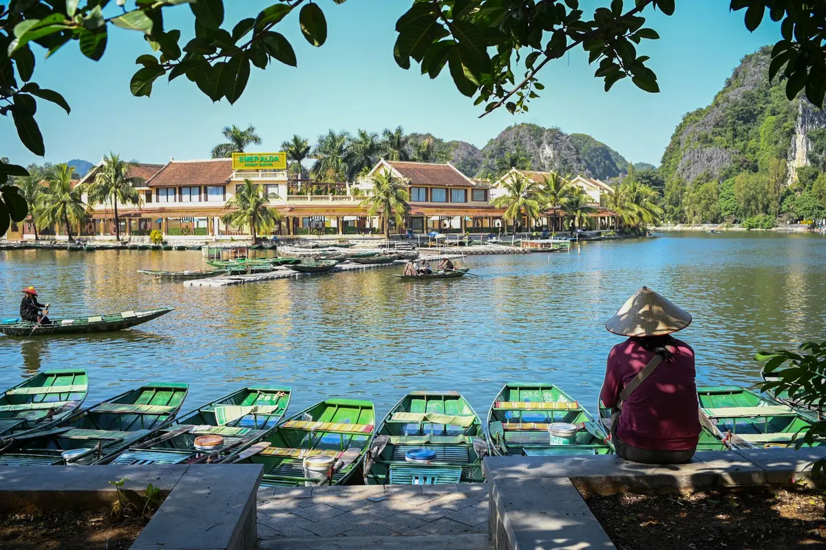 A woman in a conical hat sits beside moored green rowing boats at a river wharf in Ninh Binh while a rower crosses the water