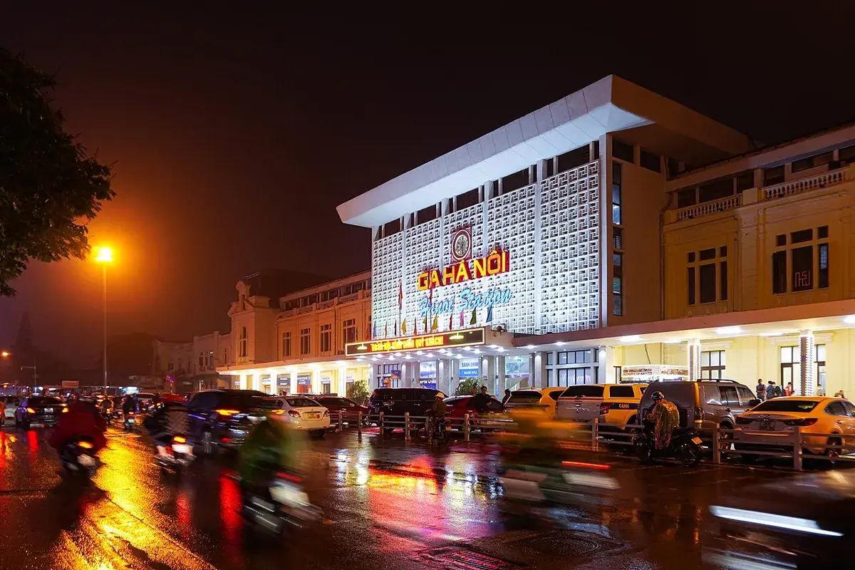 The lit front of Ga Ha Noi, Hanoi railway station, at night on a wet evening with taxis and motorbikes outside