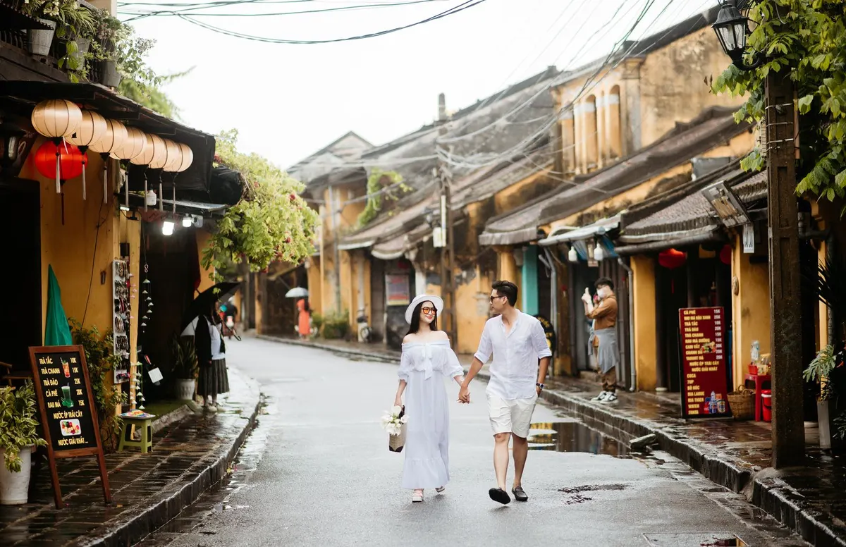 A couple holding hands walk down a rain-wet Hoi An street lined with yellow shophouses and silk lanterns