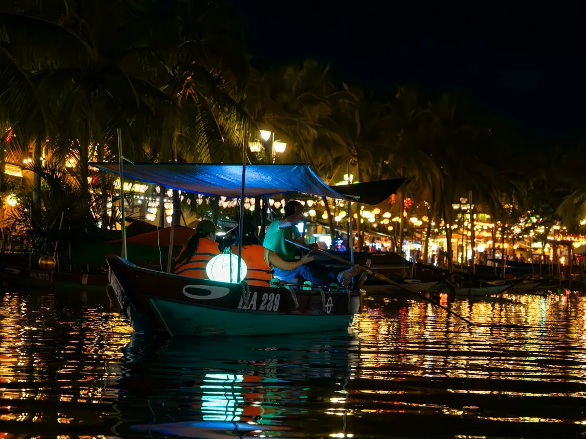 A wooden boat with a glowing lantern and a blue canopy on the Thu Bon River at night, riverside lights reflected in the water